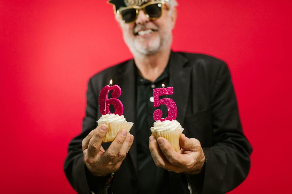 Happy senior man holding decorated cupcakes for his 65th birthday celebration against red background.
